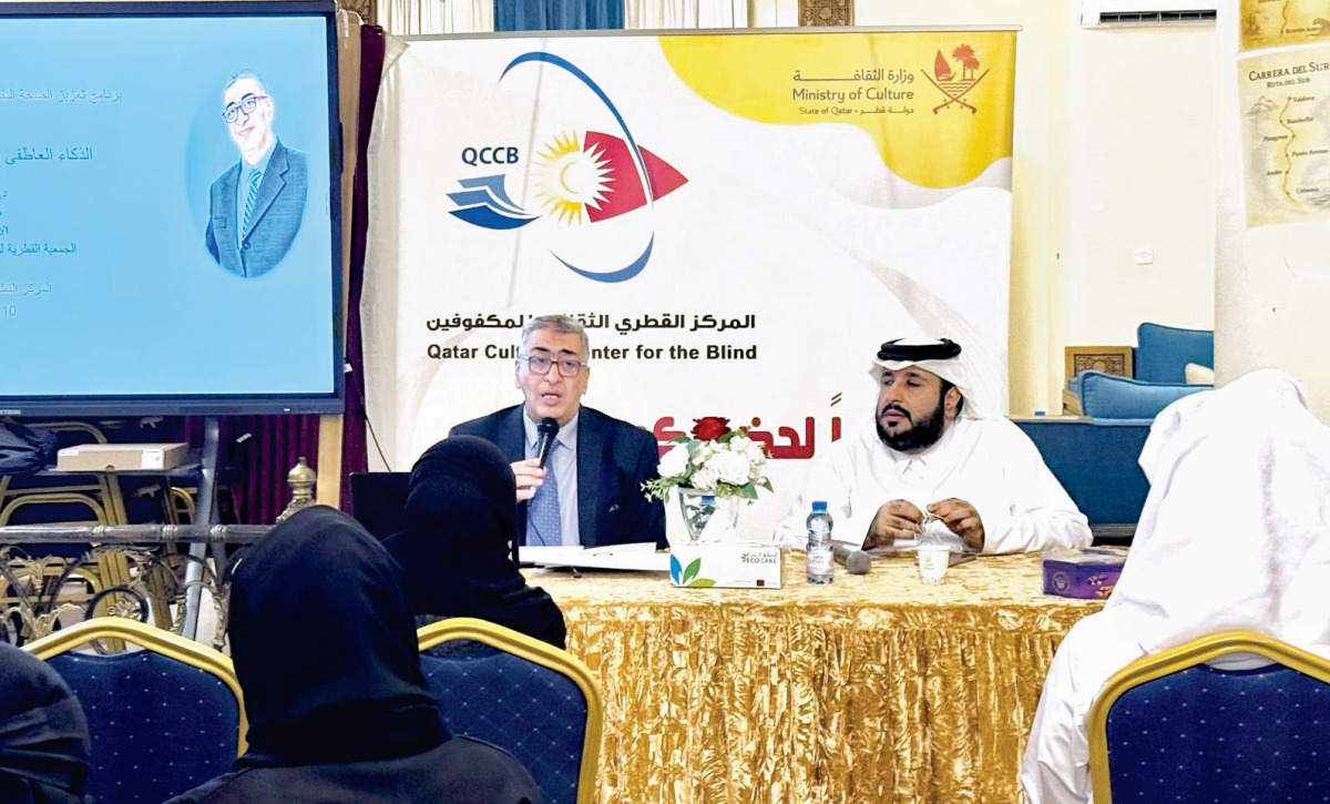 رجل يرتدي بدلة زرقاء داكنة يتحدث في ميكروفون على طاولة ذهبية اللون بجانب رجل يرتدي الثوب الأبيض والغترة، خلفهما لافتة مركز قطر الثقافي للمكفوفين وشعار وزارة الثقافة، وحضور من المستمعين في القاعة خلال محاضرة عن الذكاء العاطفي.